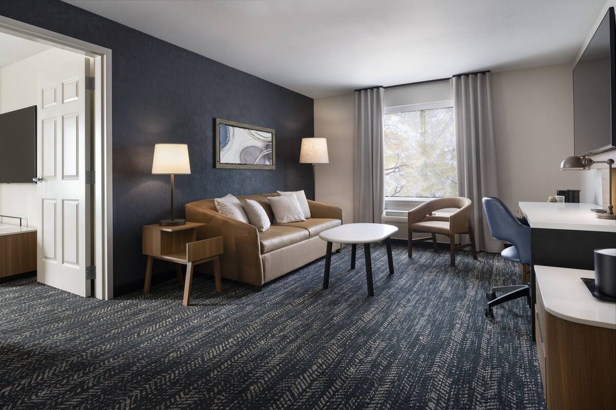 Une chambre d’hôtel avec un tapis bleu foncé, un canapé beige, une petite table ronde, un bureau avec une chaise et une grande fenêtre avec des rideaux.