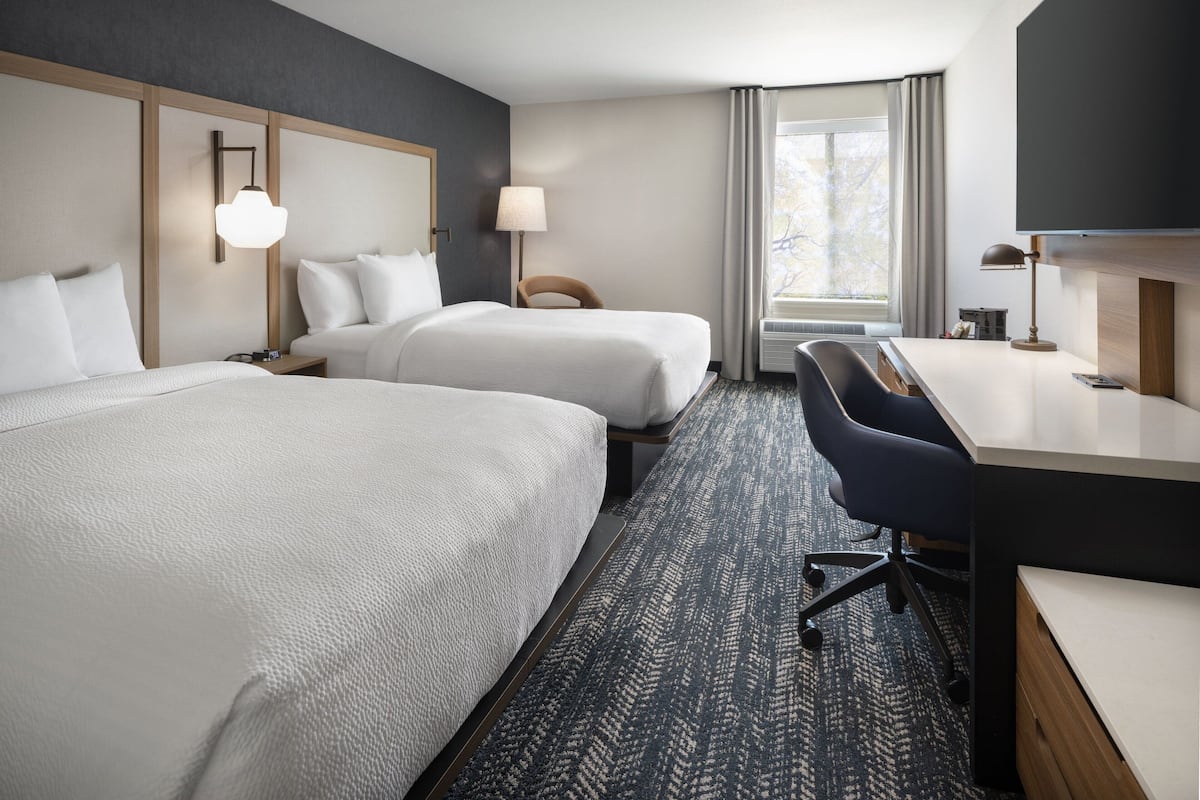 Une chambre d’hôtel avec deux lits, un bureau, une chaise, une lampe et une grande fenêtre.
