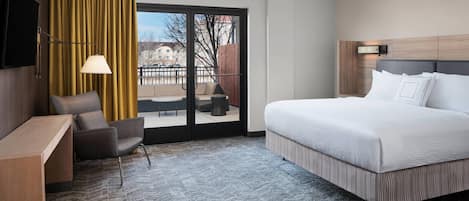 A hotel room with a large bed, a desk, a chair, a TV, and a view of the outdoors through a sliding door.