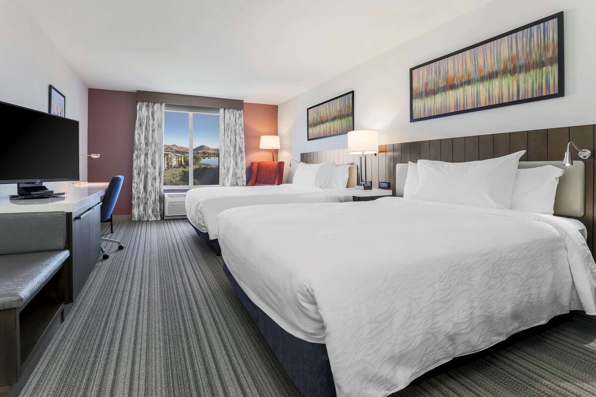A hotel room with two beds, a desk with a television, and a view of a cityscape.