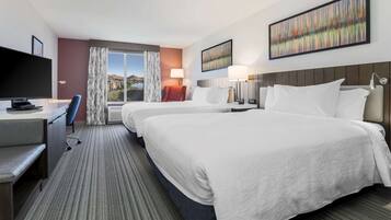 A hotel room with two beds, a desk with a television, and a view of a cityscape.