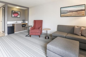 A modern living room with a grey sofa, a red armchair, and a small round table. There is a kitchen area with a microwave and a coffee machine. A painting is on the wall above the sofa.