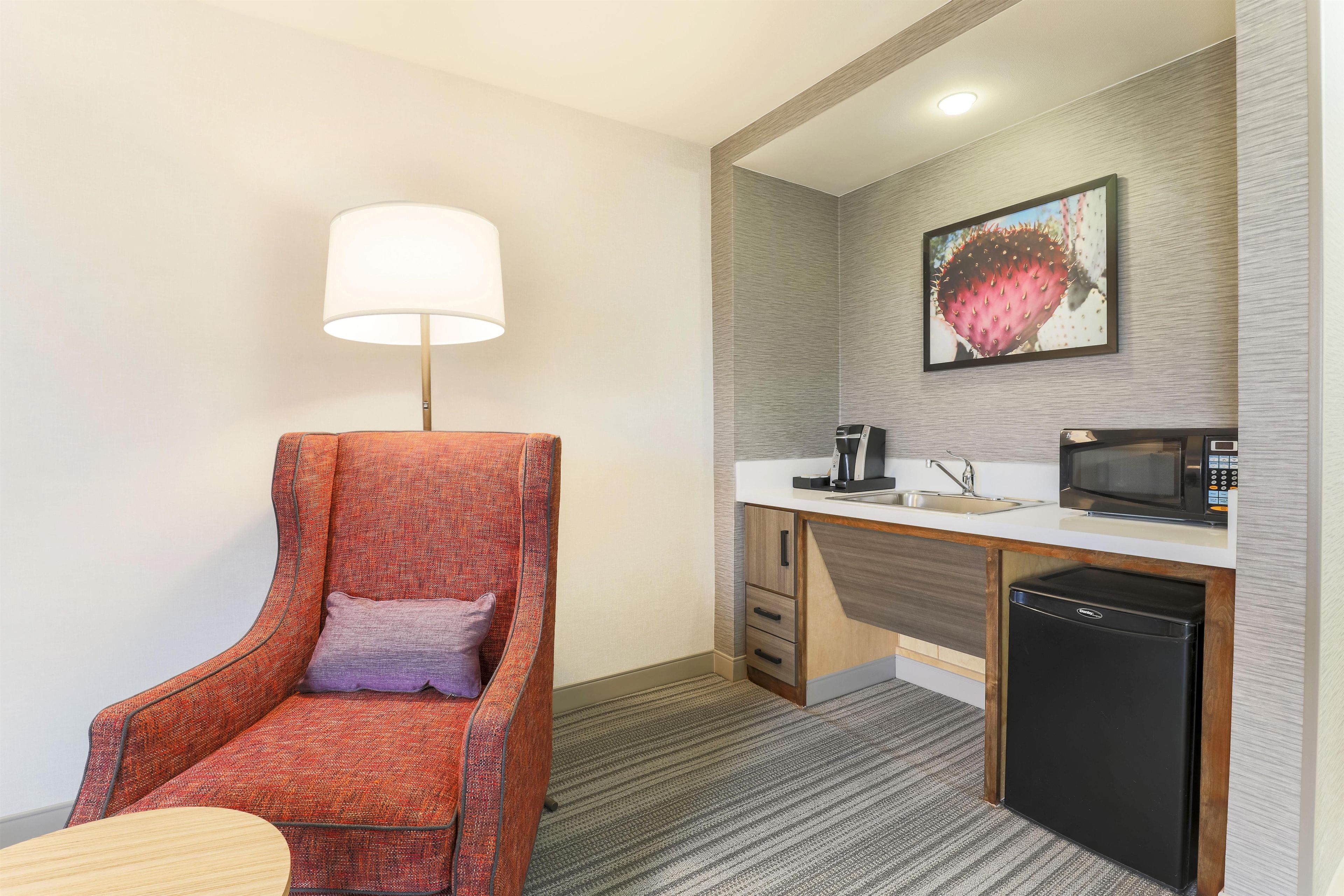A hotel room with a single orange armchair, a small table, a lamp, a kitchenette with a microwave, a coffee maker, and a refrigerator.