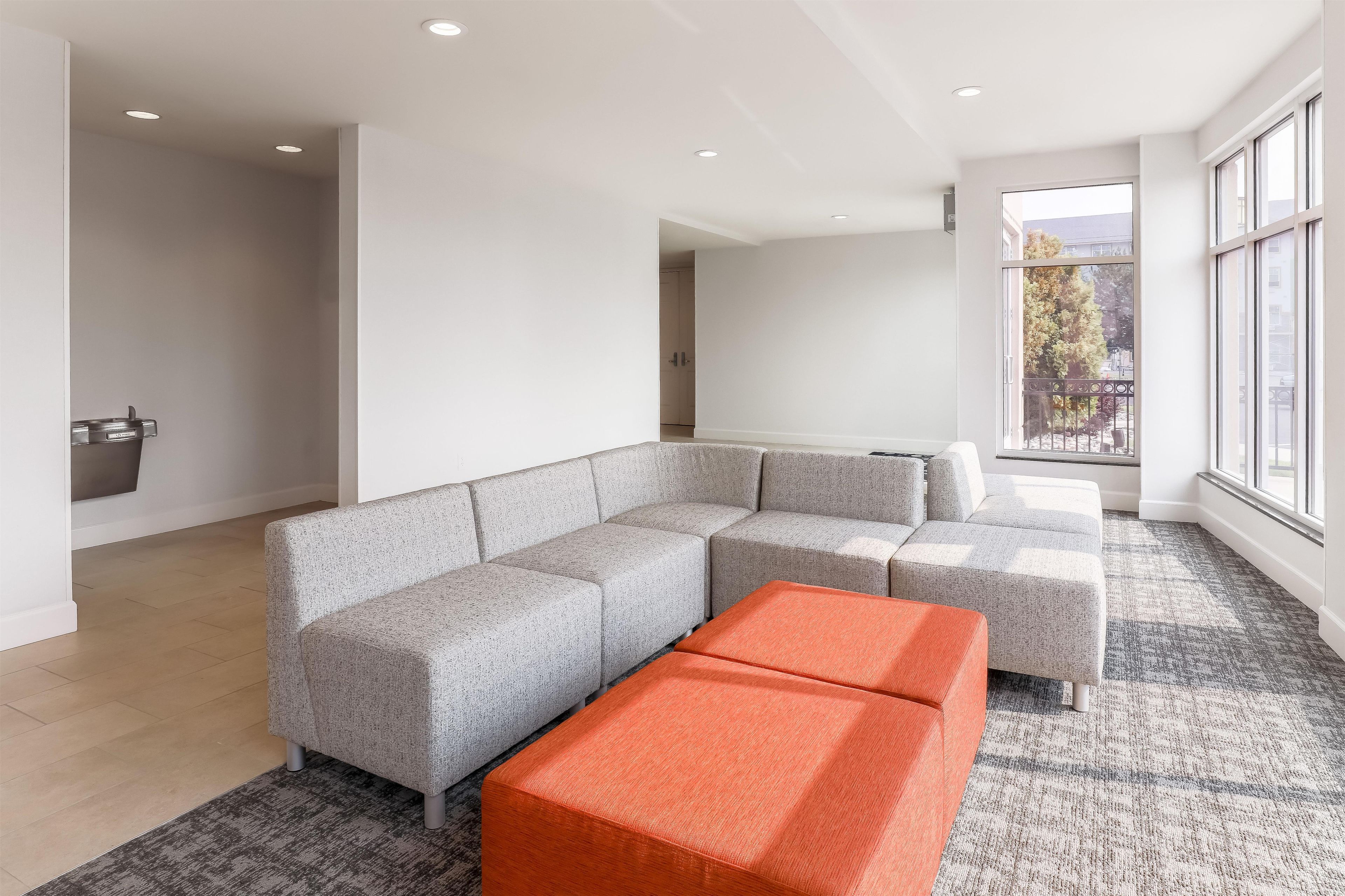 Een moderne woonkamer met een hoekbank, een helder raam en een klein oranje poef.