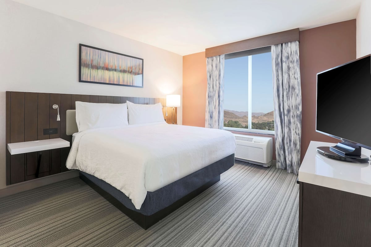 A hotel room with a large bed, a flat-screen TV, a window with curtains, and a painting on the wall.