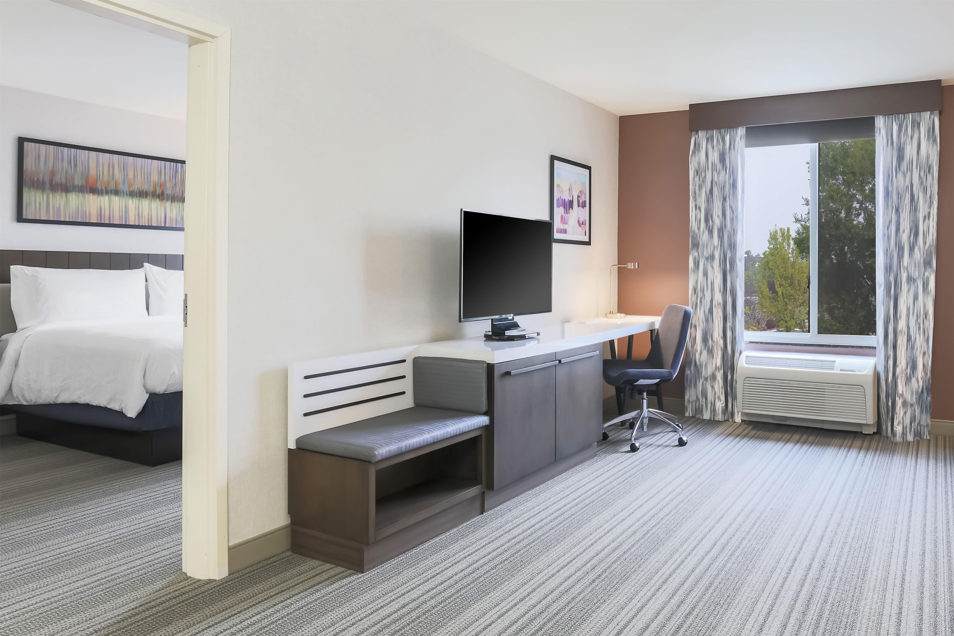 A hotel room with a bed, a desk with a computer, a chair, a TV, and a window with curtains.