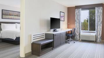 A hotel room with a bed, a desk with a computer, a chair, a TV, and a window with curtains.