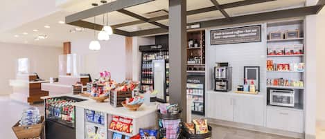 Interior de una cafetería con una barra que exhibe aperitivos, una sección refrigerada para bebidas y un letrero que indica que hay café especial disponible todo el día.