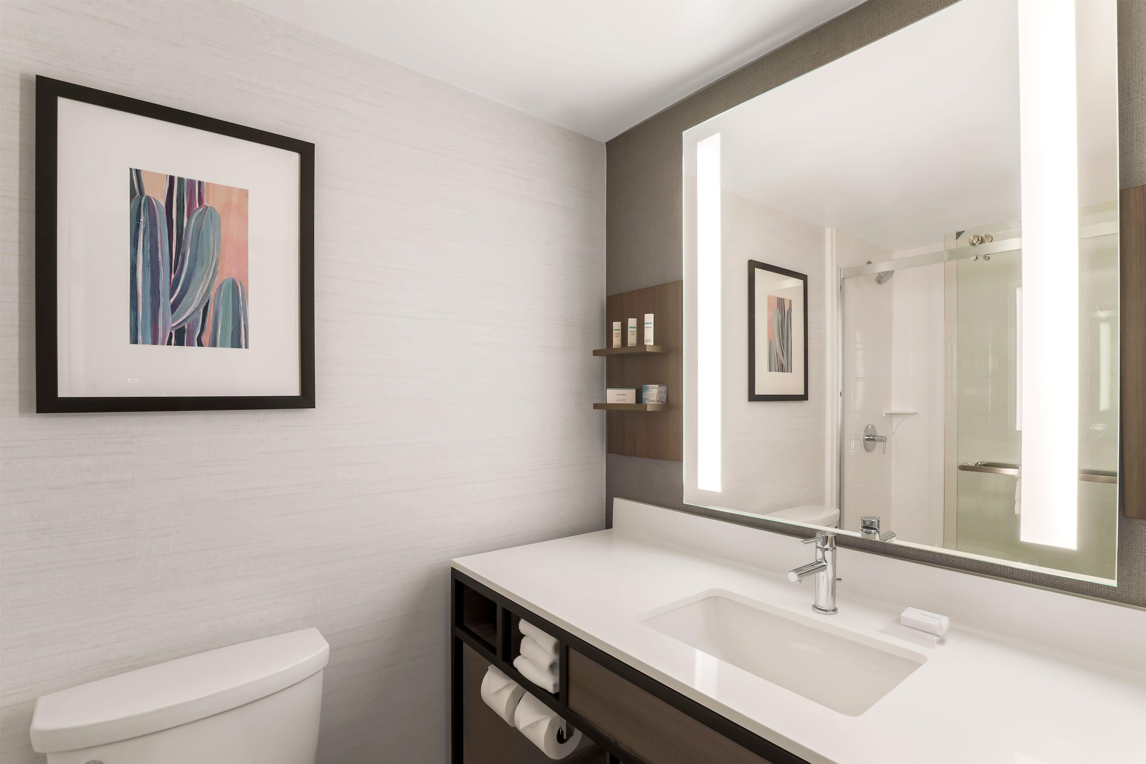 A modern bathroom with a white sink, a large mirror, and a framed abstract painting on the wall.