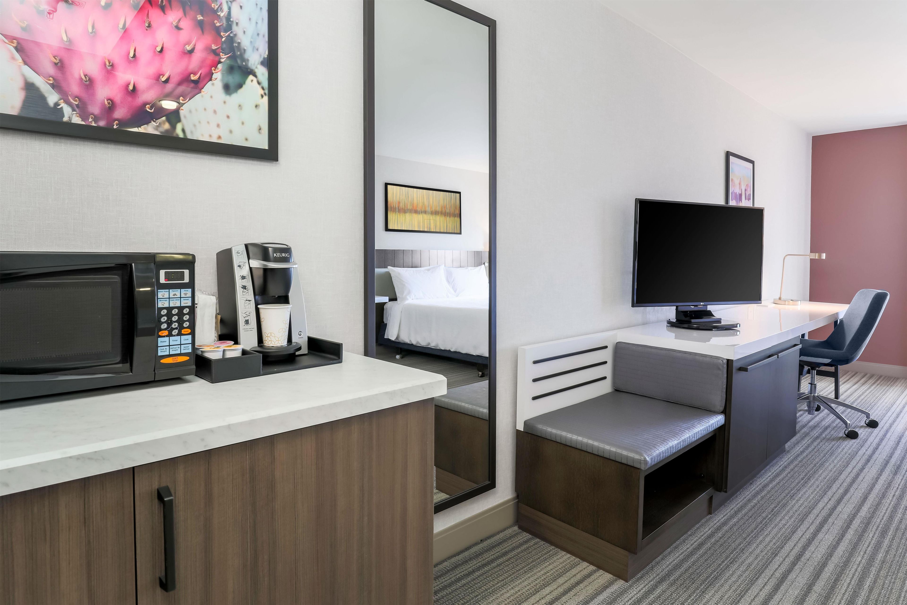 A hotel room with a kitchenette, a bed, a desk, a chair, a television, and a large mirror.