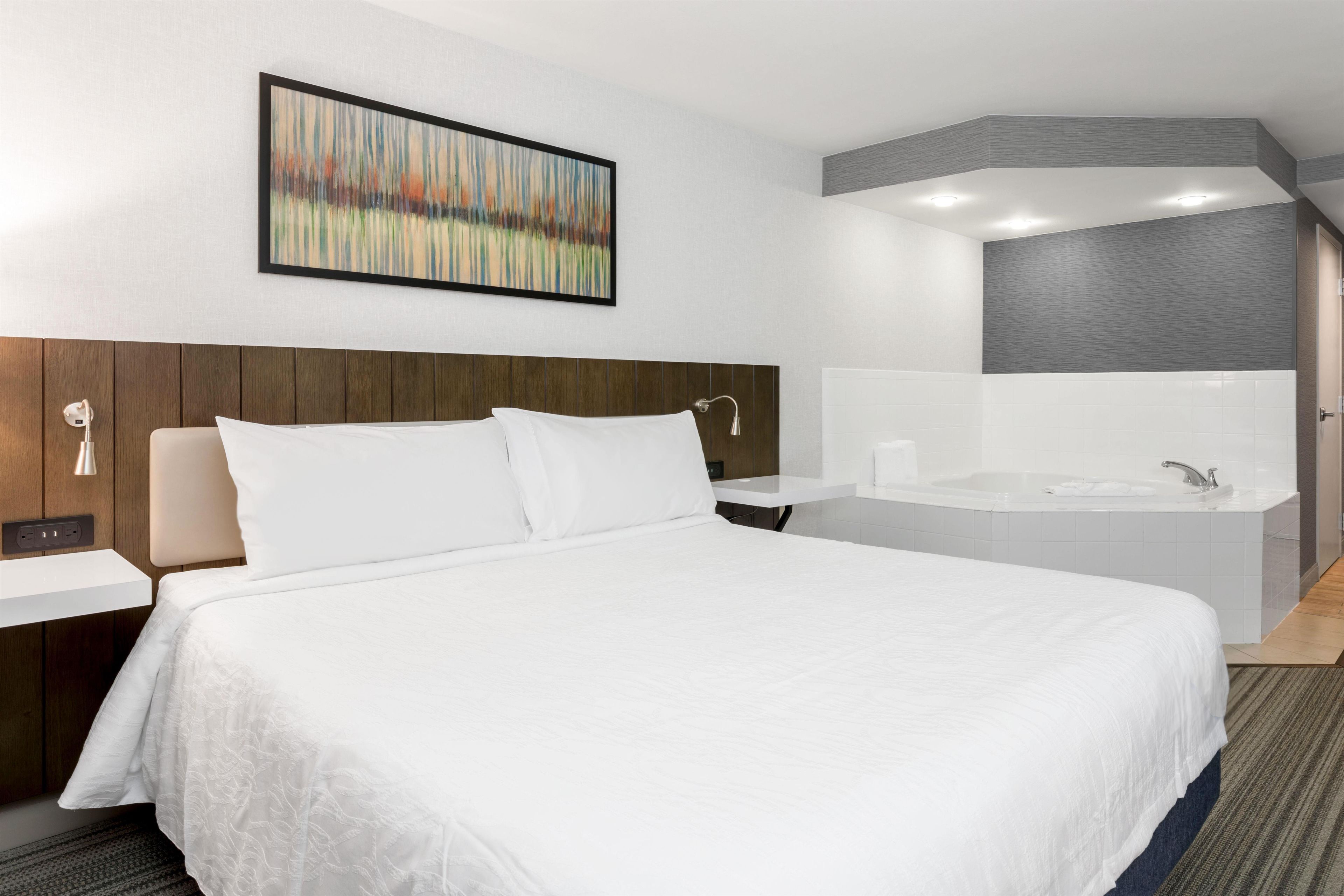 Hotellihuone, jossa on suuri sänky, yöpöytä, seinälle ripustettu taulu sekä kylpyhuone, jossa on kylpyamme ja pesuallas.