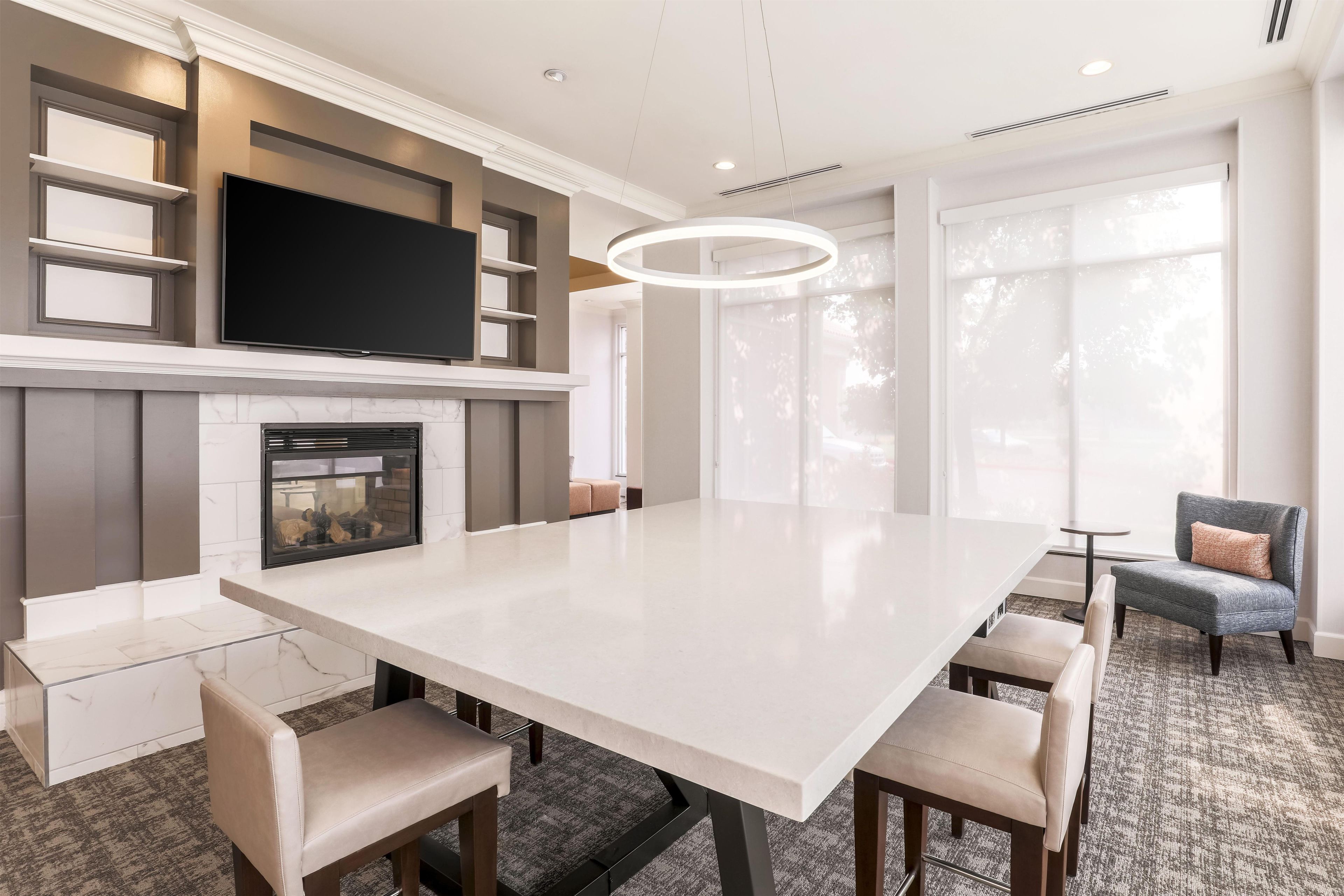 A modern dining room with a large white table, multiple chairs, a fireplace, and a television mounted on the wall.