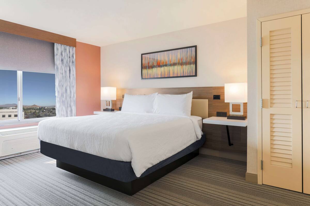 A hotel room with a large bed, two bedside tables, a nightstand, a window with a view, and a door.