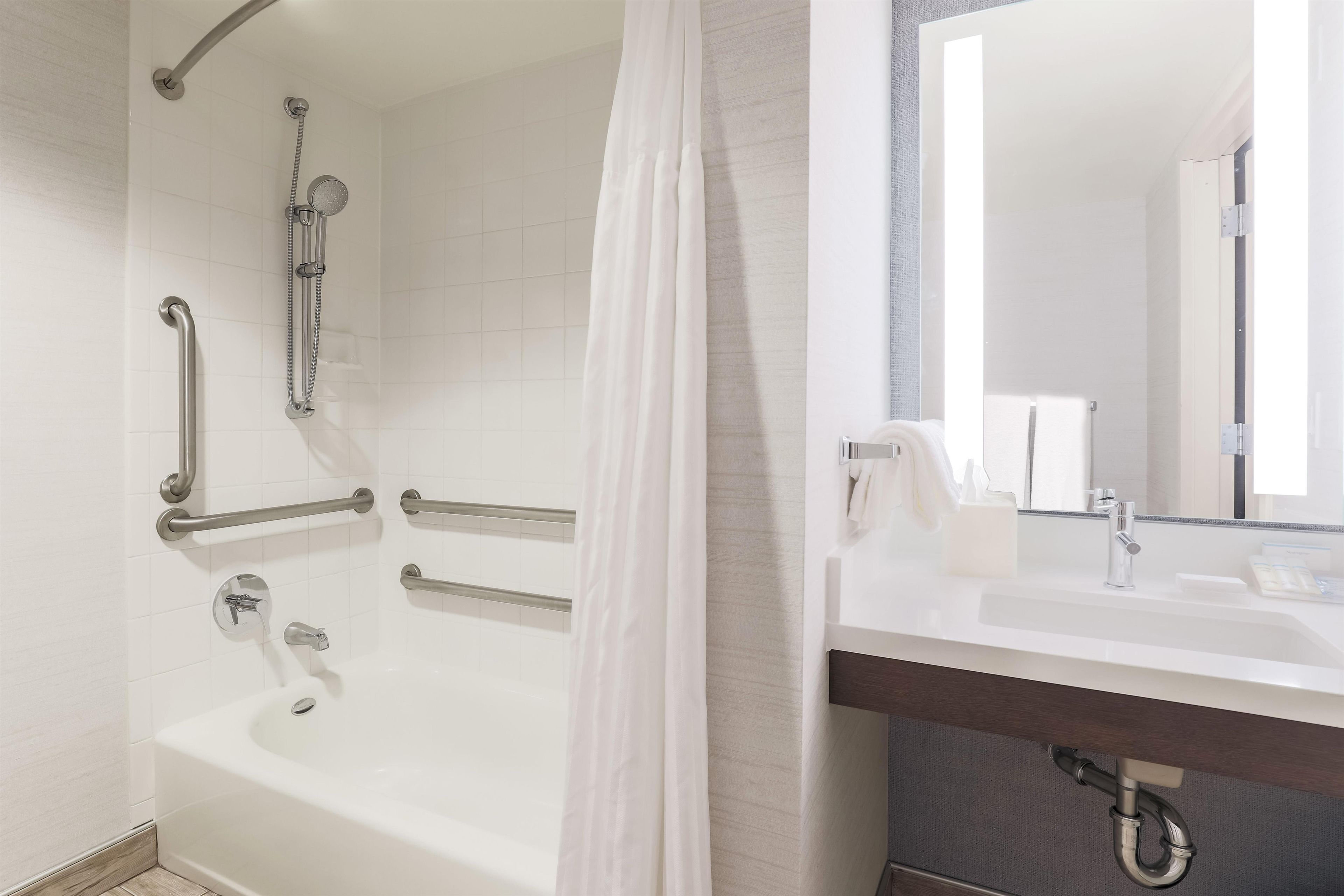 Ein Badezimmer mit einer Badewanne, einer Duschkabine inklusive Haltegriffen, einem Waschbecken mit Spiegel und einem Handtuchhalter.