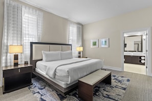 Une chambre d’hôtel avec un grand lit, des tables de chevet, un banc, une salle de bain visible à travers une porte ouverte et un tapis à motifs.