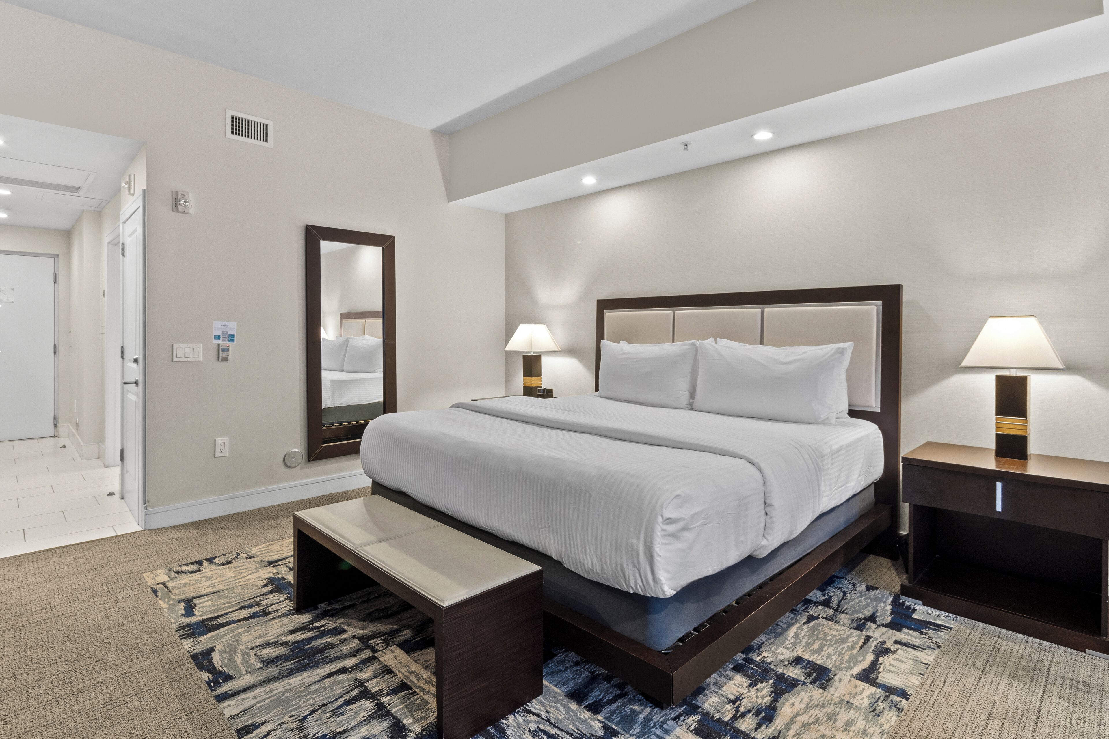 A hotel room with a large bed, bedside lamps, a bench, and a patterned rug.