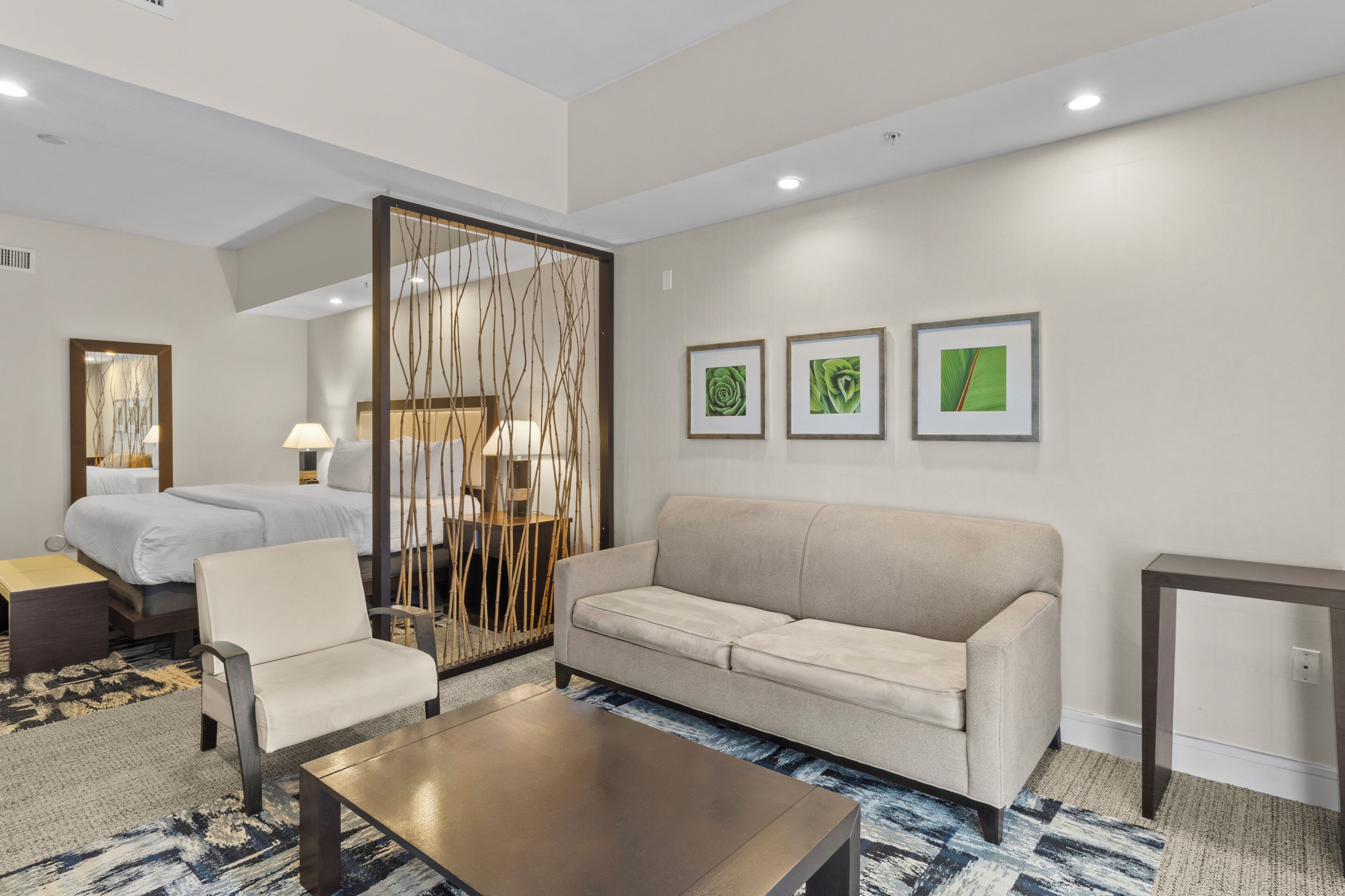 Une chambre d’hôtel comprenant un lit, un canapé, une chaise, une table basse, un bureau et un paravent décoratif. Trois tableaux encadrés ornent le mur.