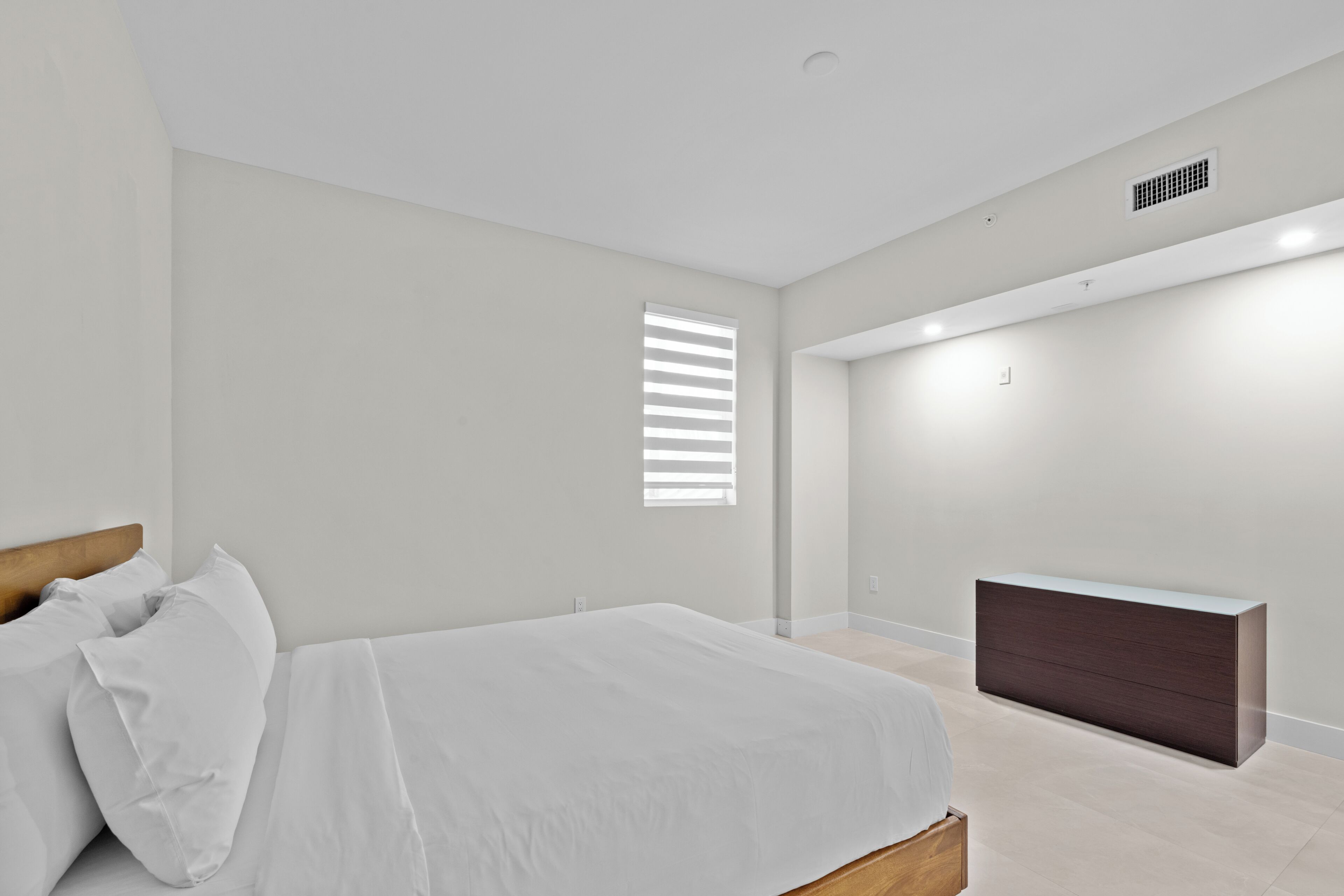 A bedroom with a bed, a dresser, a window with blinds, and a ceiling vent.