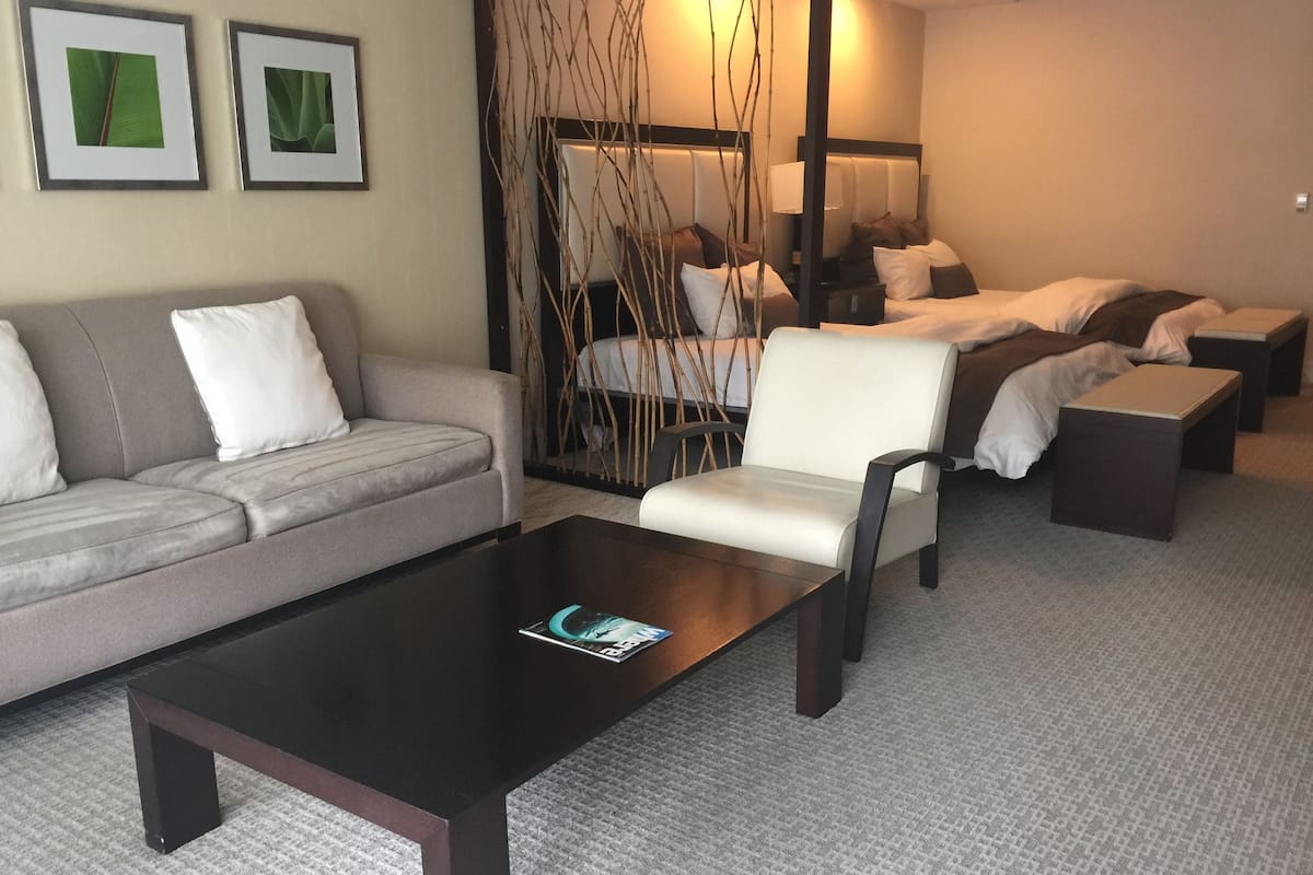 Une chambre d’hôtel moderne avec un canapé, un fauteuil et une petite table basse. On y trouve deux lits avec une literie blanche et un panneau décoratif au mur.