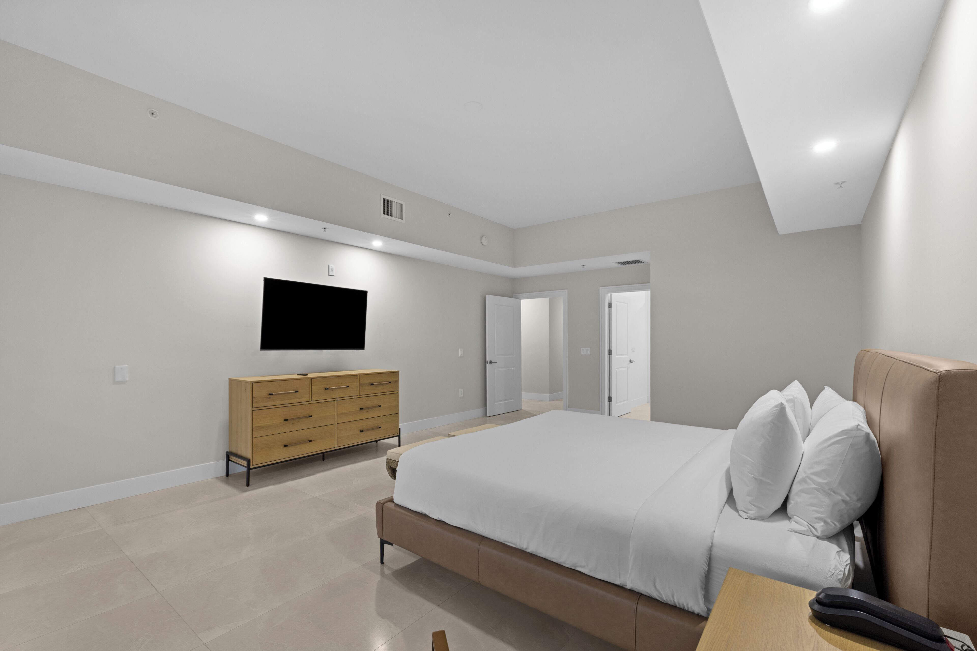A modern bedroom with a large bed, a wooden dresser, a flat-screen TV, and a door leading to another room.