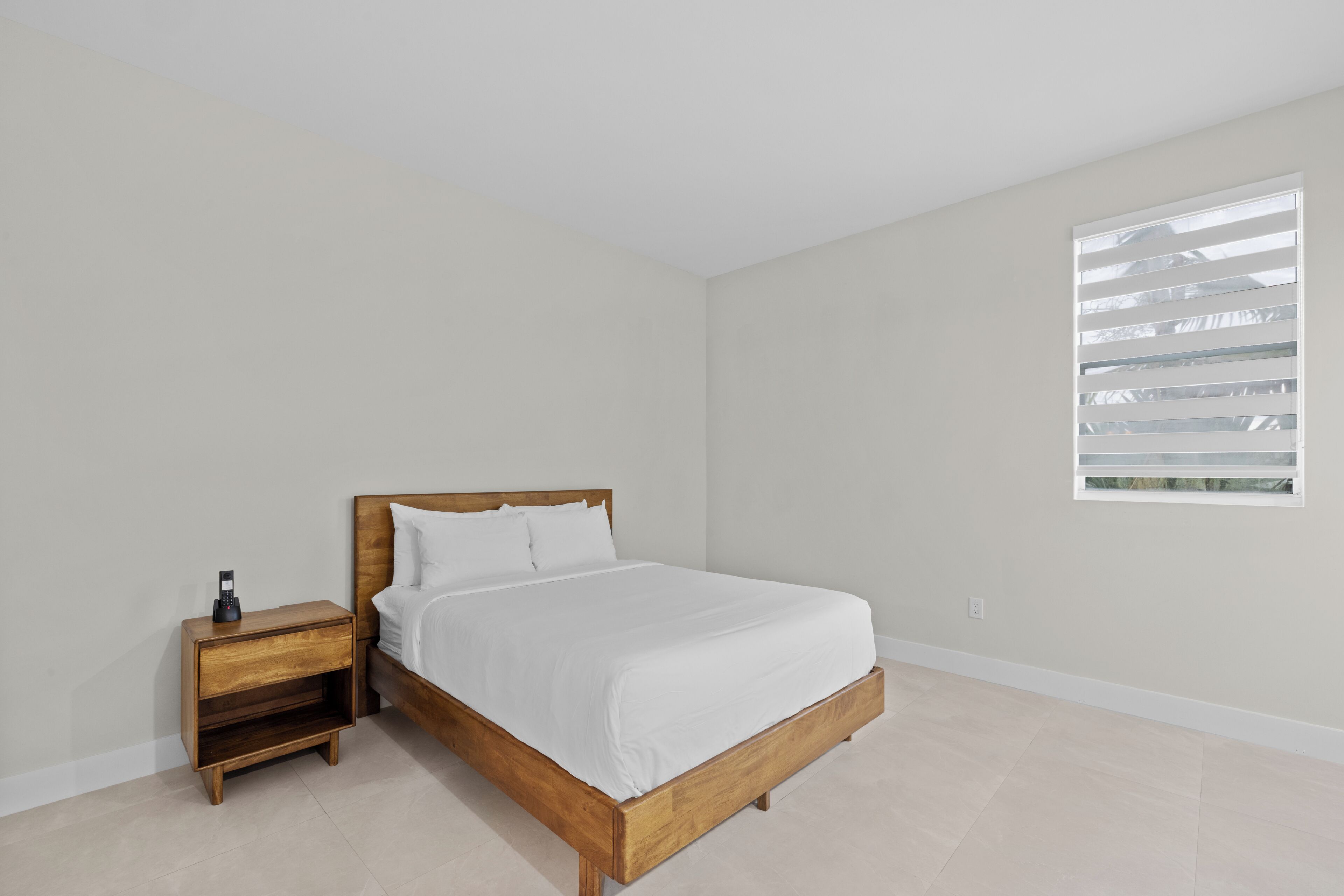 Un dormitorio con cama de madera, ropa de cama blanca, una mesita de noche con teléfono y una ventana con persianas.