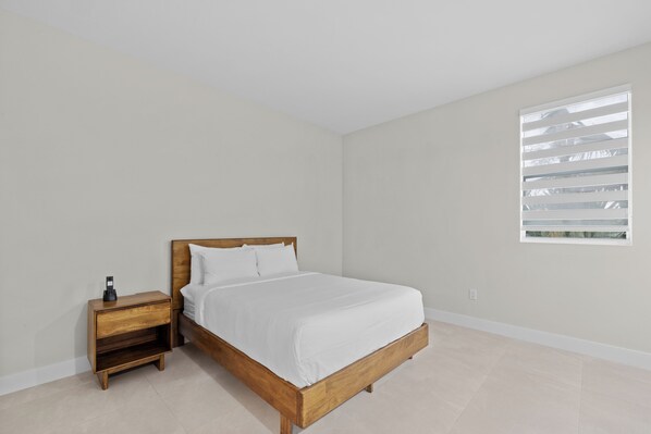A bedroom with a wooden bed, white bedding, a nightstand with a phone, and a window with blinds.