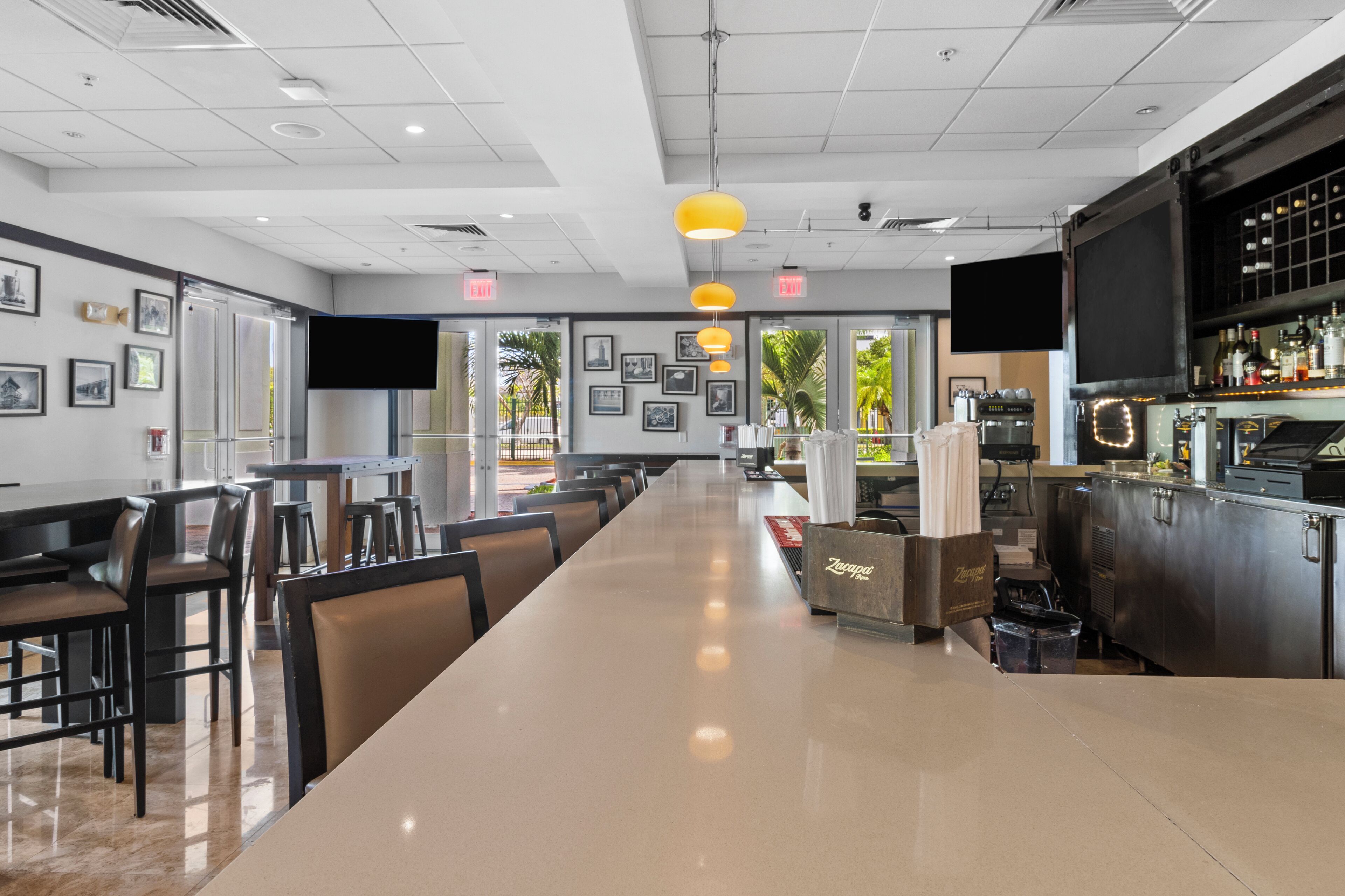 A modern restaurant with a long counter, multiple tables, and chairs. There are framed pictures on the walls, a bar area with bottles, and a large screen.
