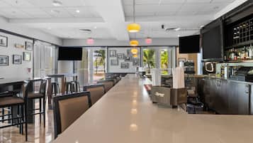 A modern restaurant with a long counter, multiple tables, and chairs. There are framed pictures on the walls, a bar area with bottles, and a large screen.