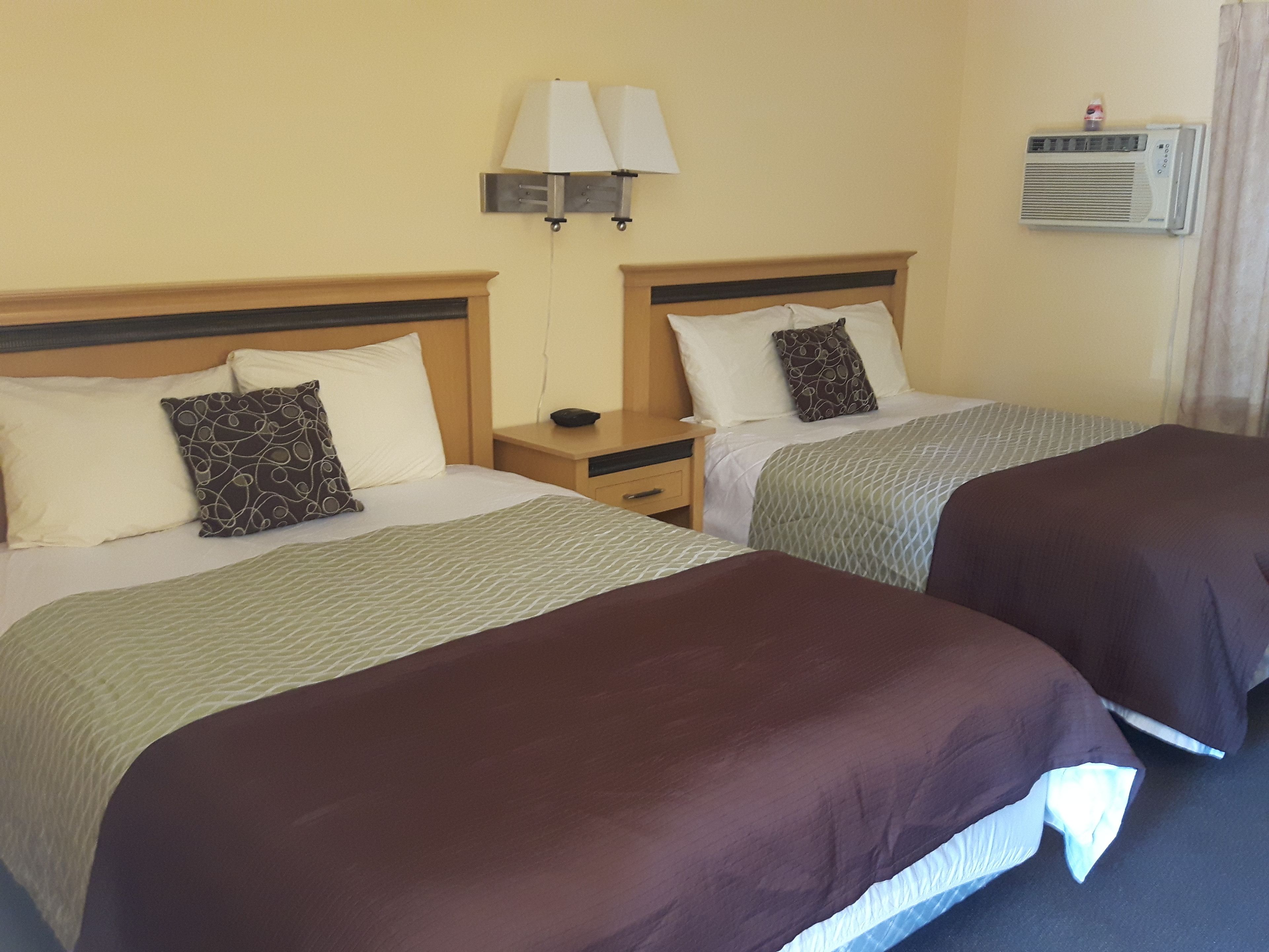 A hotel room with two beds, each with a headboard, a bedside table, a lamp, and an air conditioning unit.
