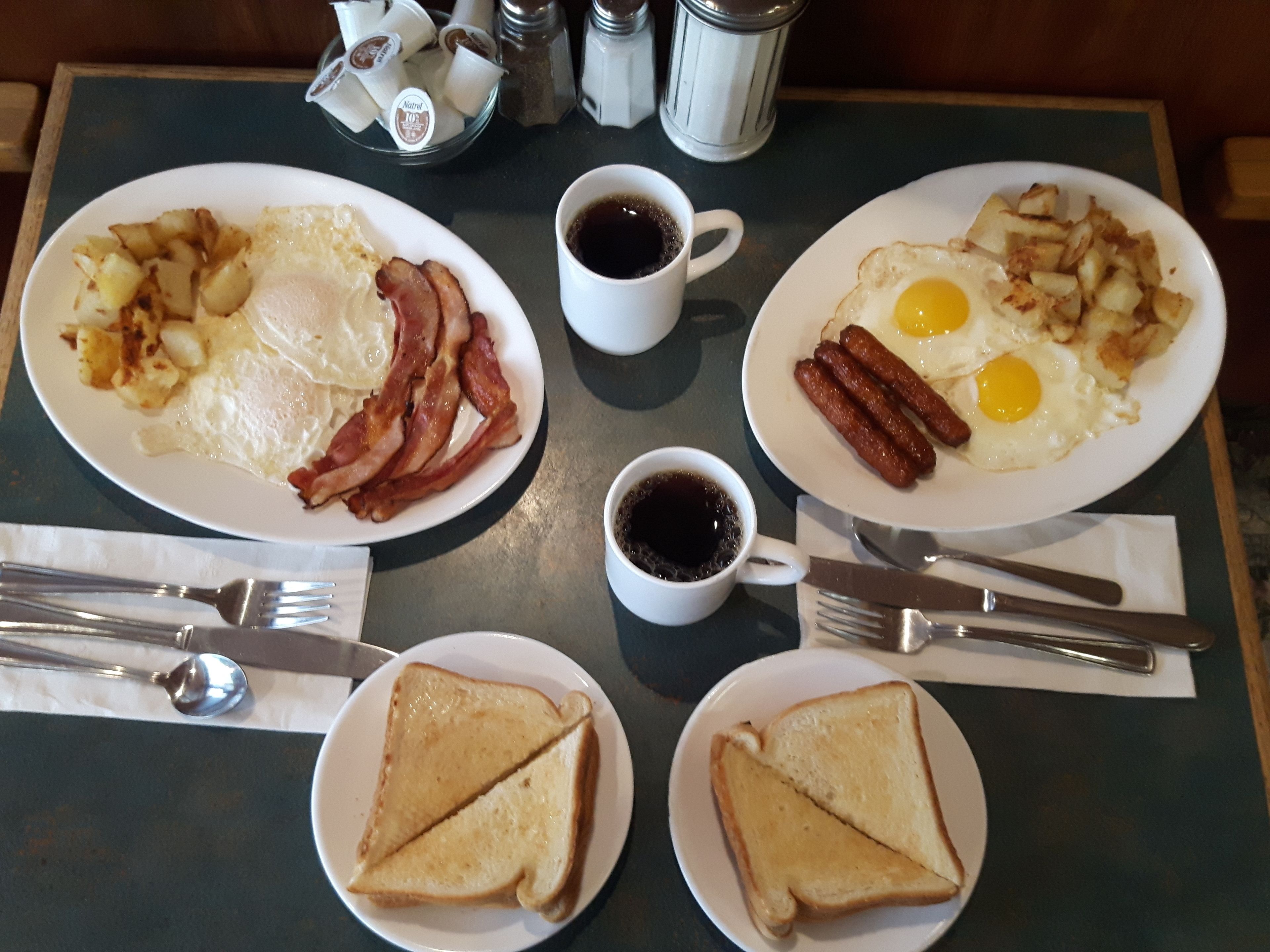 A breakfast setting with plates of eggs, bacon, potatoes, and toast, along with coffee cups and cutlery on a table.