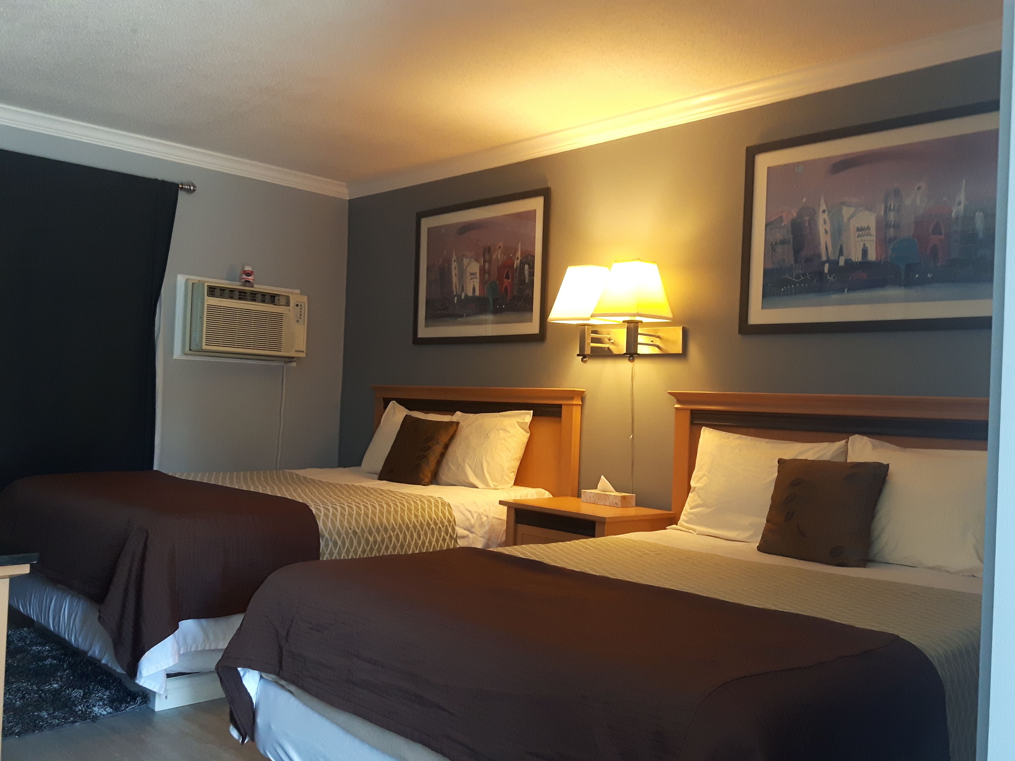 Una habitación de hotel con dos camas, una mesita de noche, una lámpara, un cuadro y un aire acondicionado instalado en la pared.