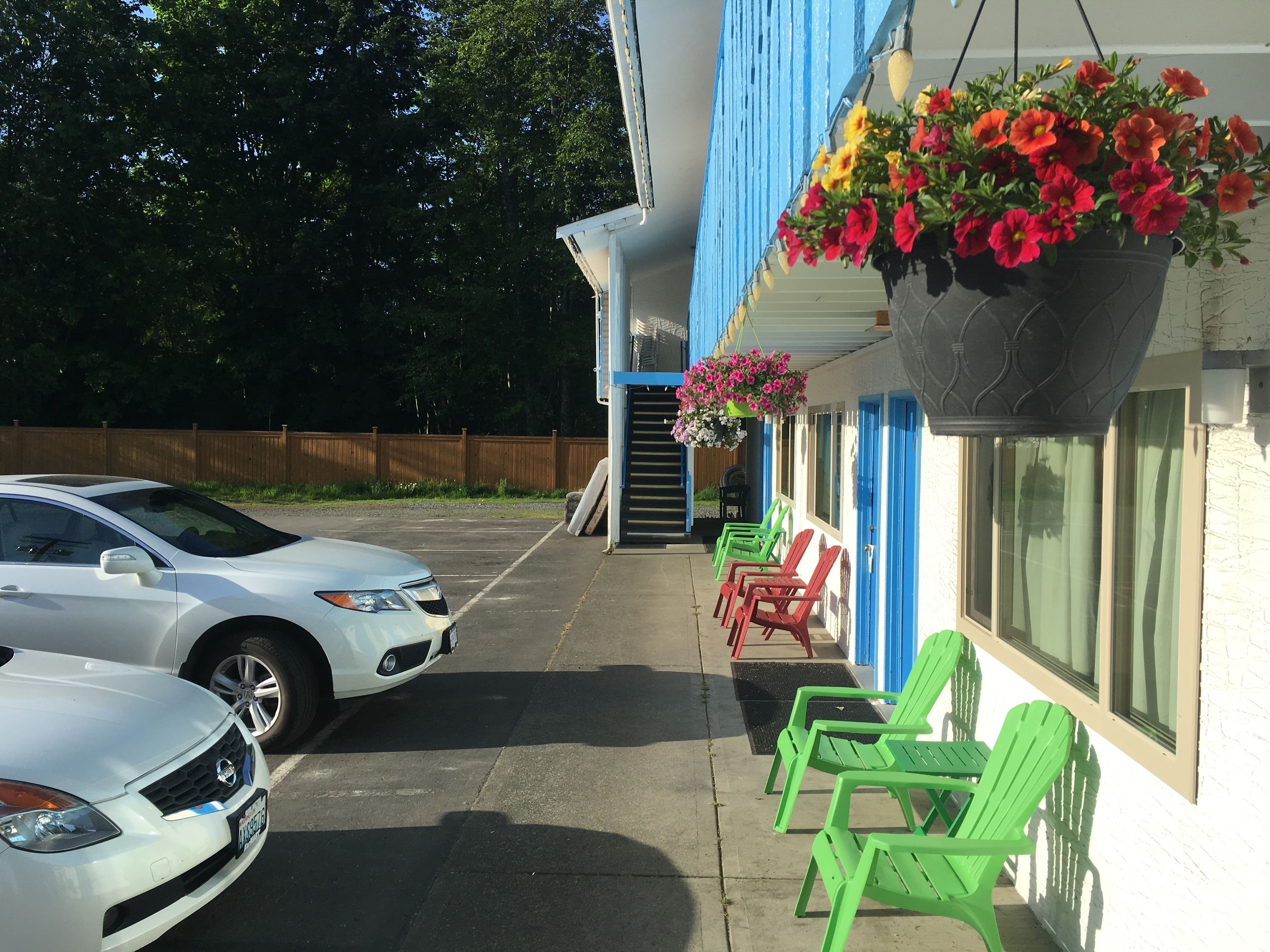 A parking area with a white car and a green chair in front of a building with blue trim and a hanging flower basket.