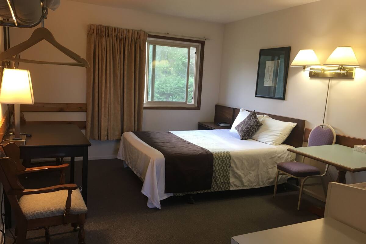 A hotel room with a bed, bedside table, desk, chair, and a window with curtains.