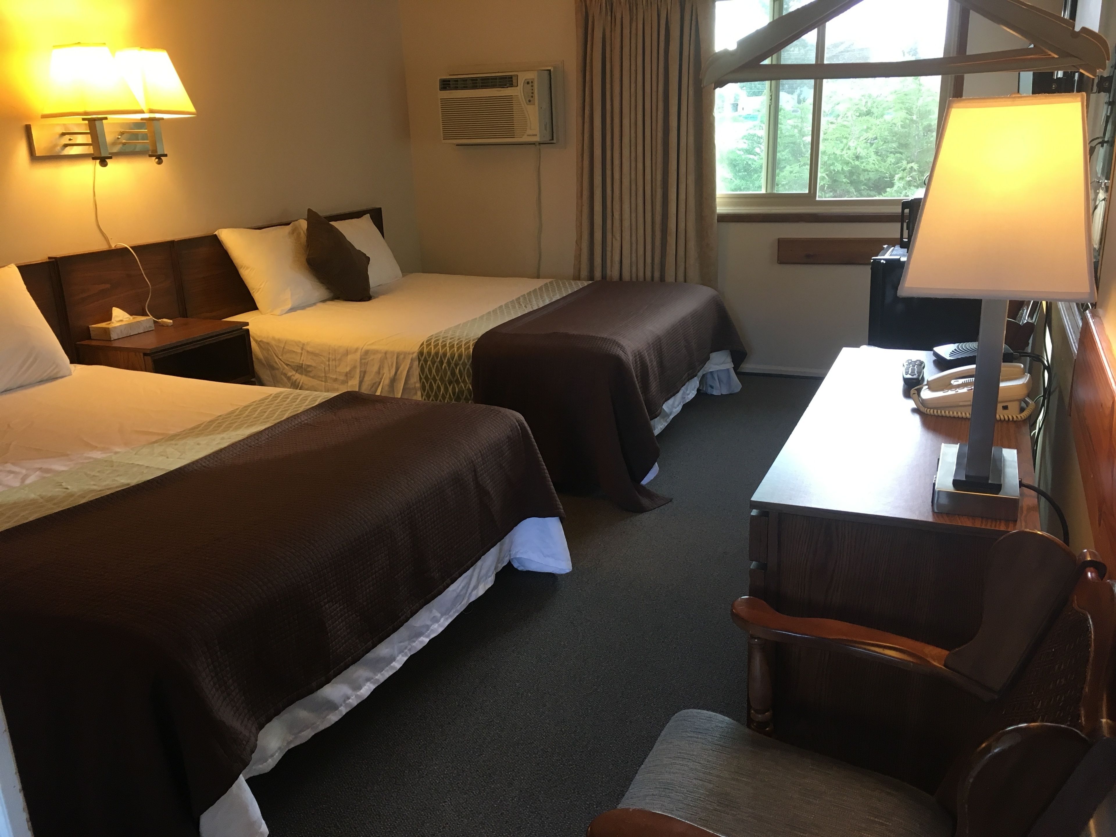 A hotel room with two beds, a desk, a chair, an air conditioning unit, and a window with curtains.