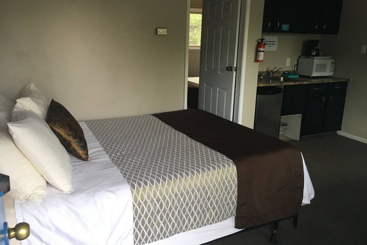 A bedroom with a bed, pillows, a door, a kitchenette with a microwave, and a fire extinguisher.