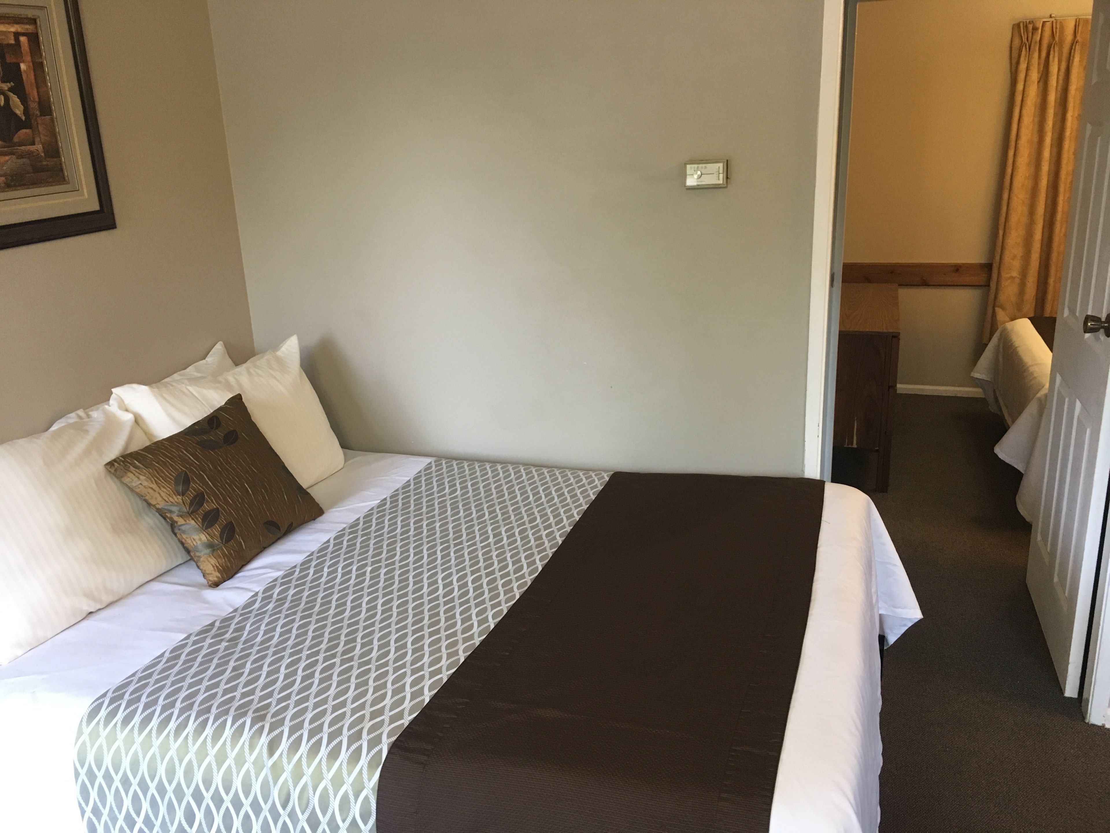 A hotel room with a bed, pillows, a thermostat, and a door leading to another room.