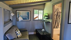 A modern living room with a grey couch, a small table, a painting, and a window with a view of greenery. - Fuller Lake Motel (Chemainus)
