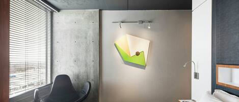 A modern hotel room with a bed, a chair, a desk, and a wall-mounted light fixture.