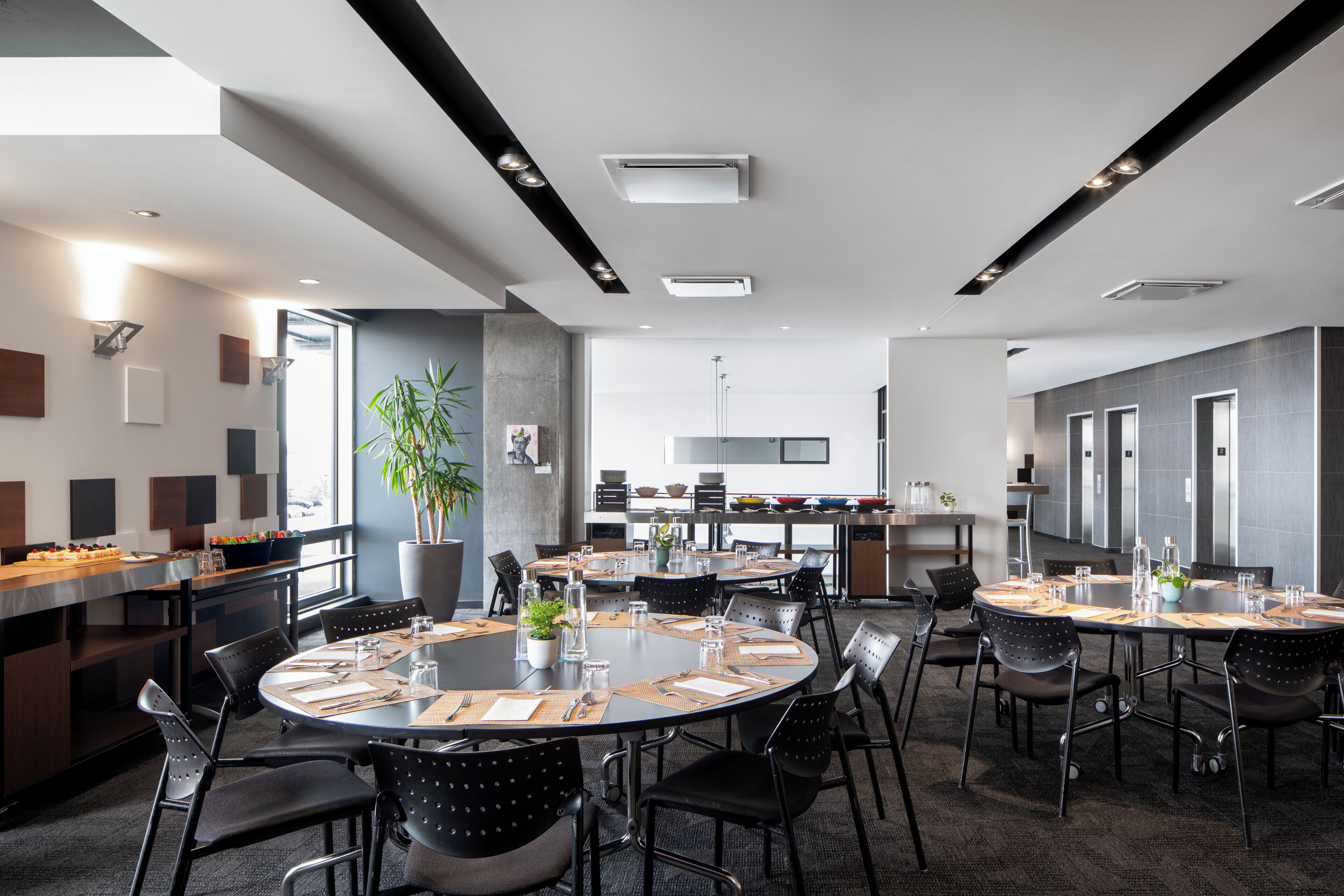 Une salle de conférence moderne, dotée de tables rondes, de chaises noires et d’un coin cuisine à l’arrière-plan.
