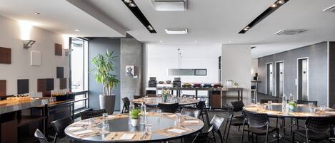 Una sala de conferencias moderna con mesas redondas, sillas negras y una cocina al fondo.