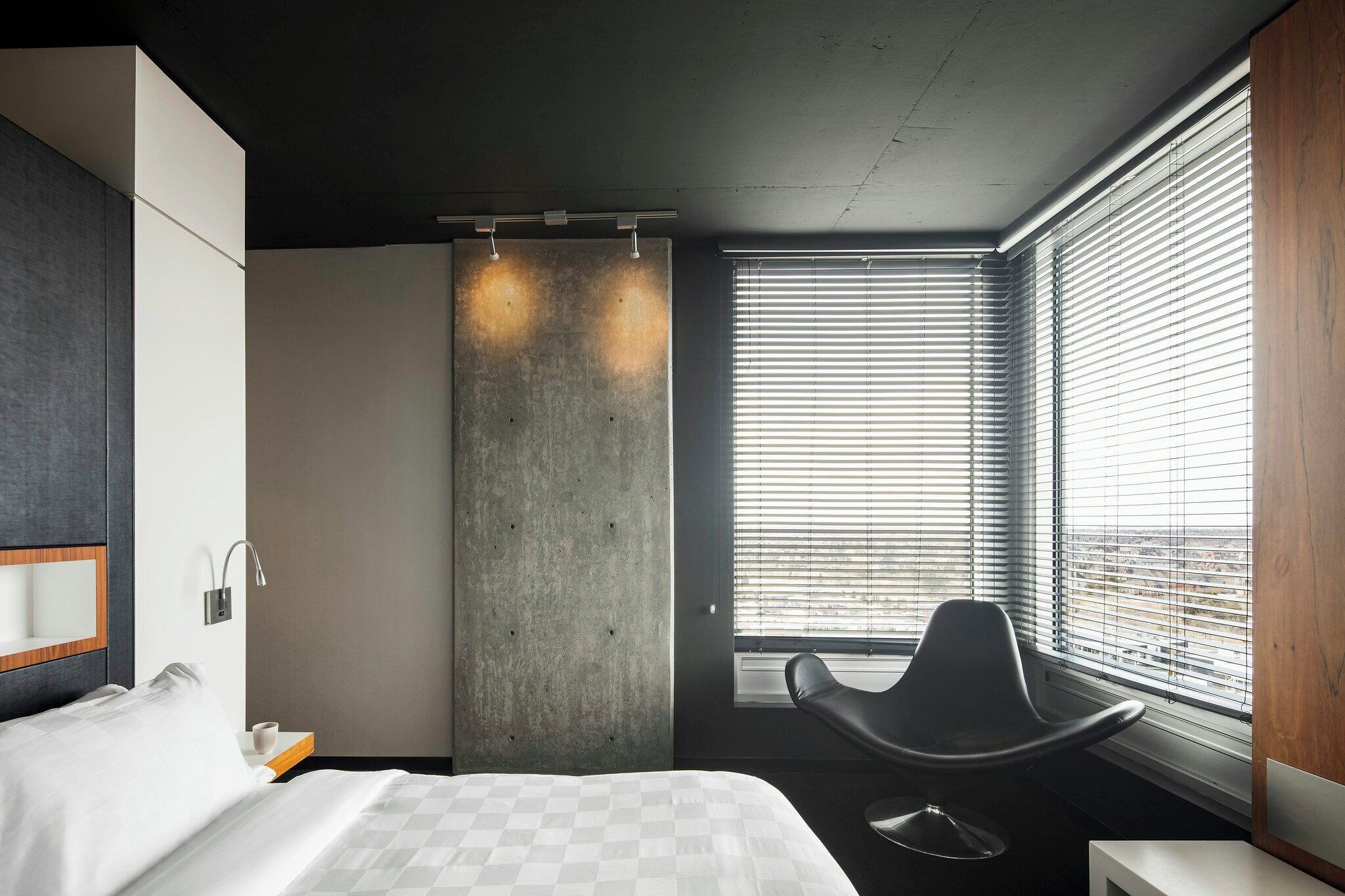 A modern hotel room with a large bed, a seating area, a concrete accent wall, and a window with blinds.