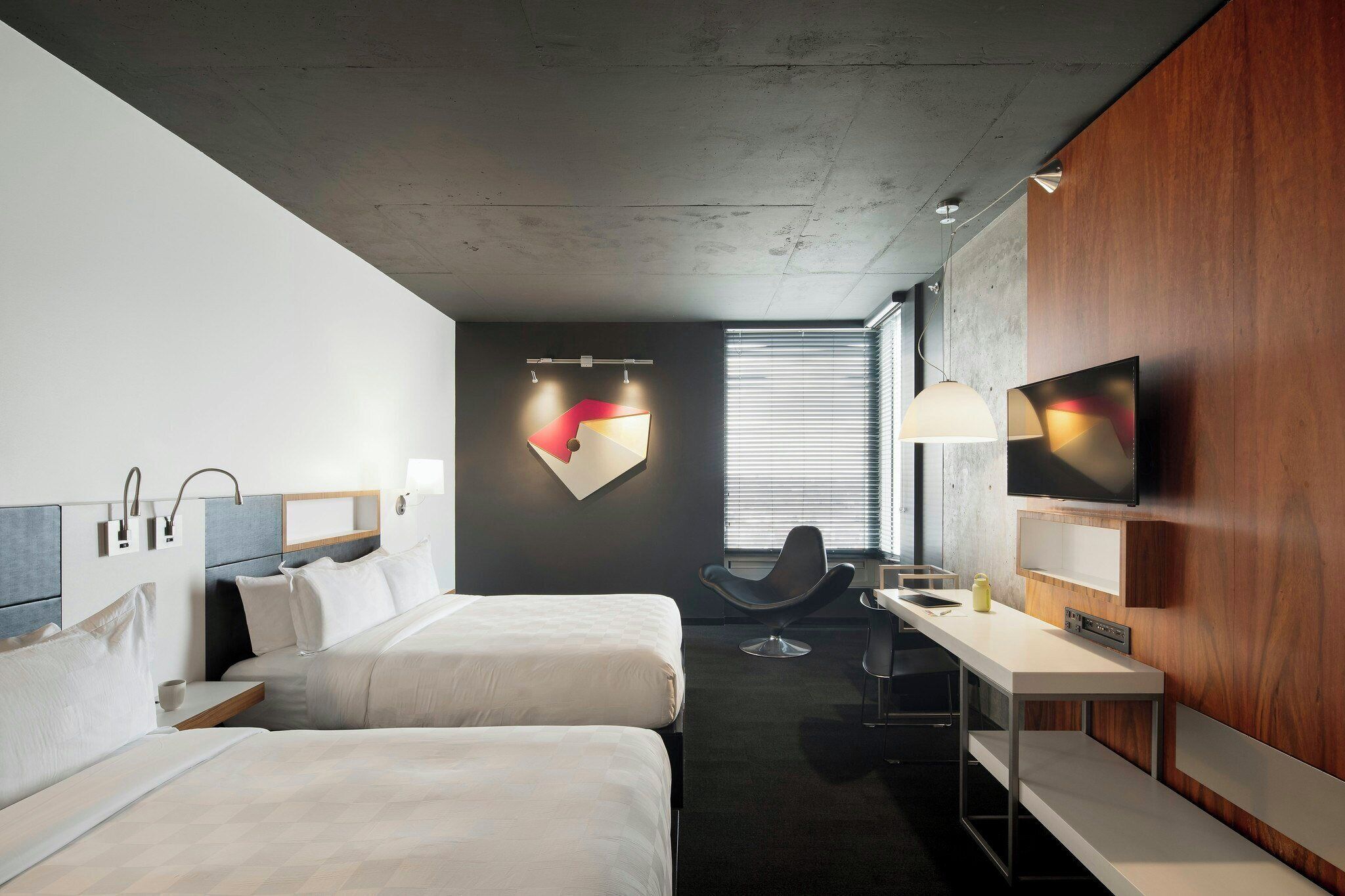 A modern hotel room with two beds, a desk, a chair, a TV, and a unique wall-mounted light fixture.