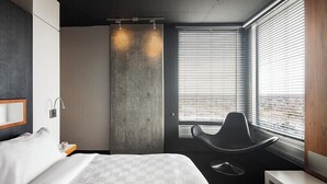 A modern hotel room with a bed, a chair, a large window with blinds, and a concrete accent wall. - Hôtel Alt Quartier DIX30 (Brossard)
