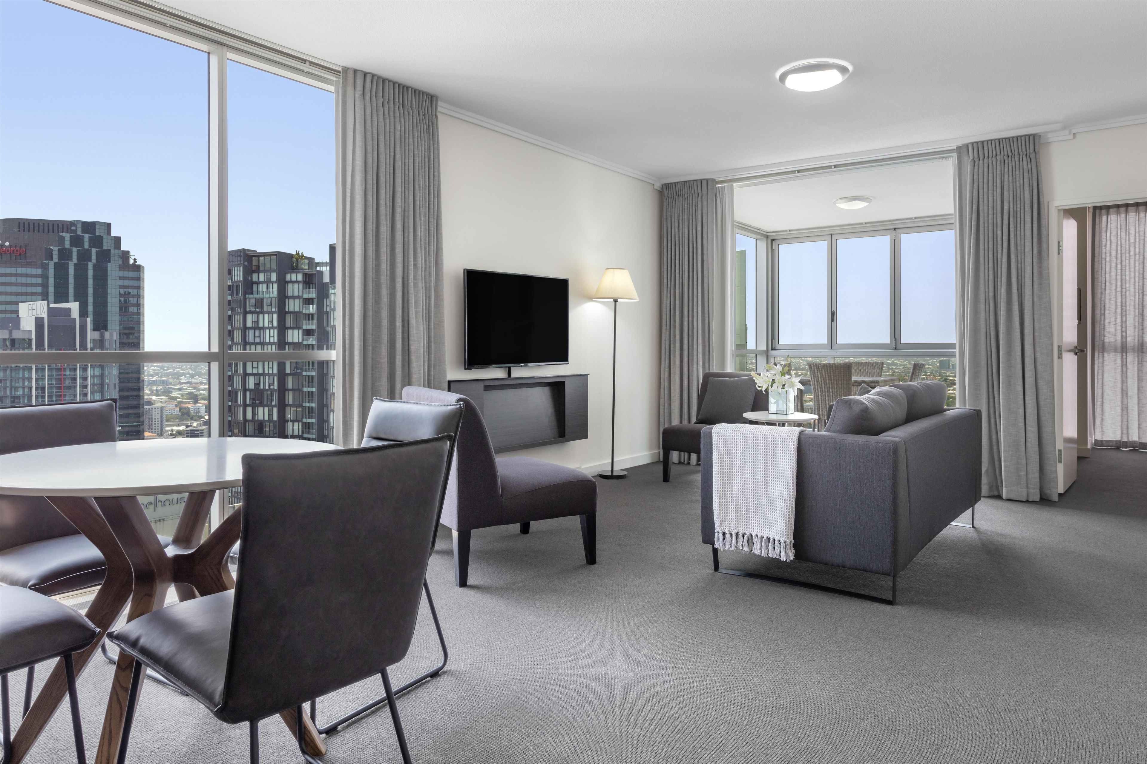 A modern hotel room with a city view, a flat-screen TV, a dining area with a table and chairs, and a seating area with a sofa and armchairs.