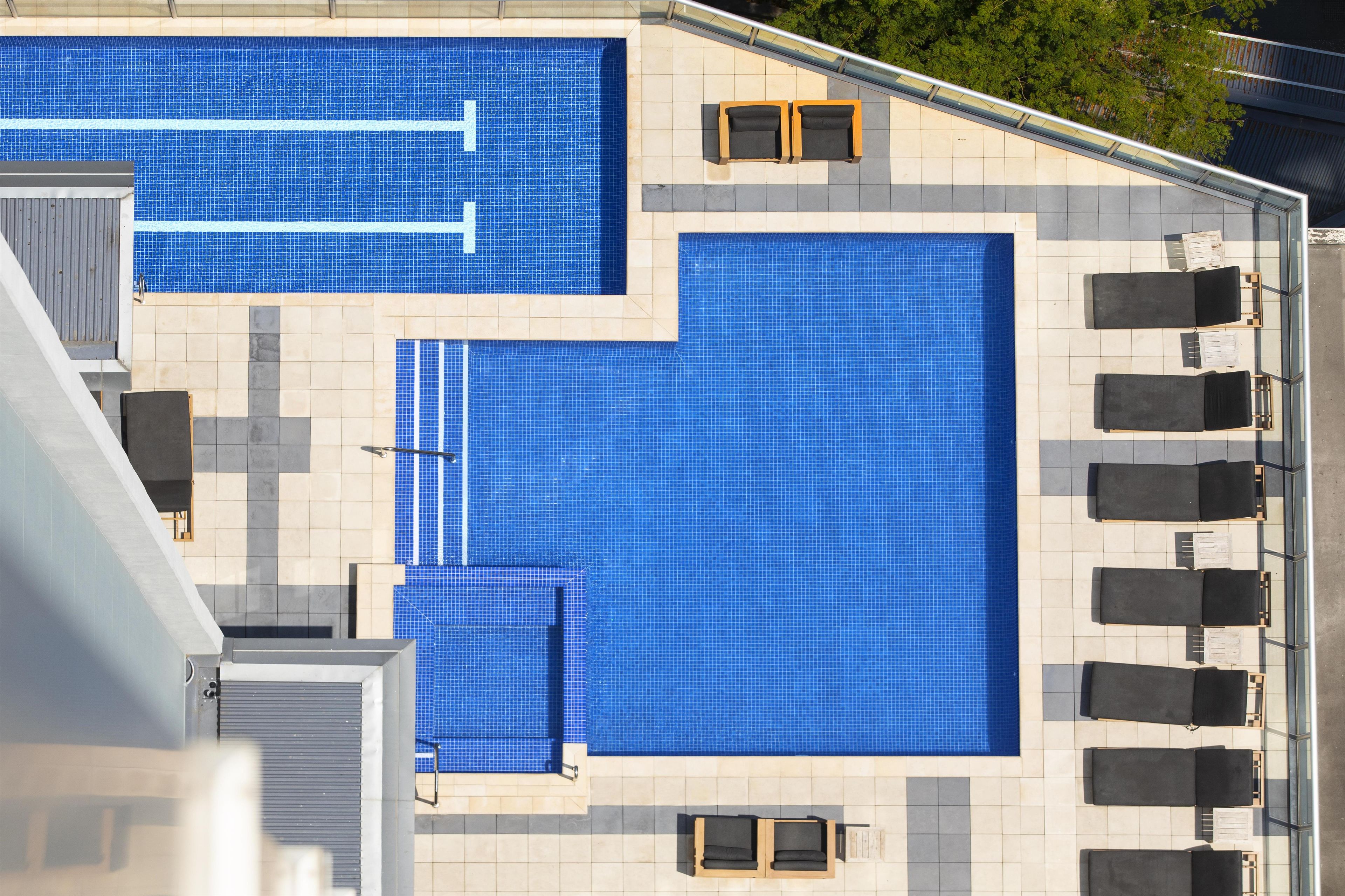 Une piscine sur le toit, avec un sol carrelé de carreaux bleus, entourée de chaises longues et protégée par une rambarde en verre.