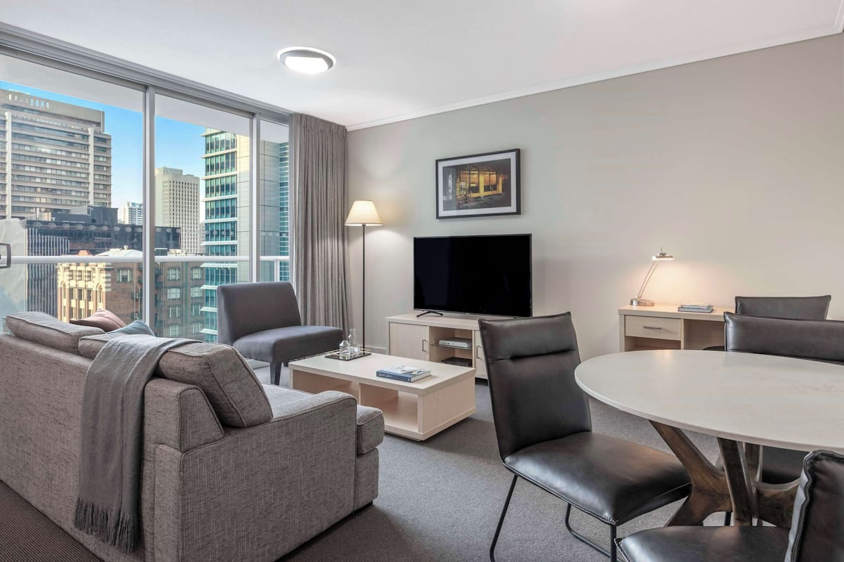 Ein modernes Hotelzimmer mit großem Fenster, Flachbildfernseher, Sofa, Couchtisch, Esstisch mit Stühlen und einer Lampe.