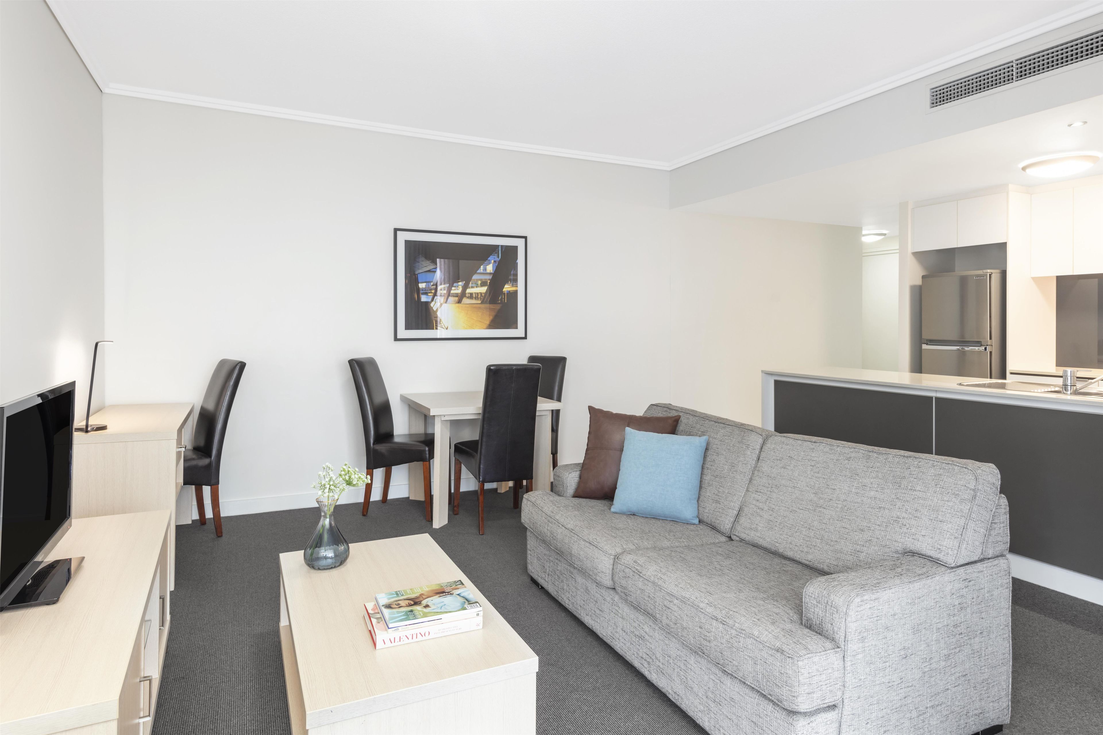 Una habitación de hotel moderna con un sofá gris, una mesa de comedor con sillas, un televisor de pantalla plana, una pequeña mesa de centro con una revista y un cuadro en la pared.