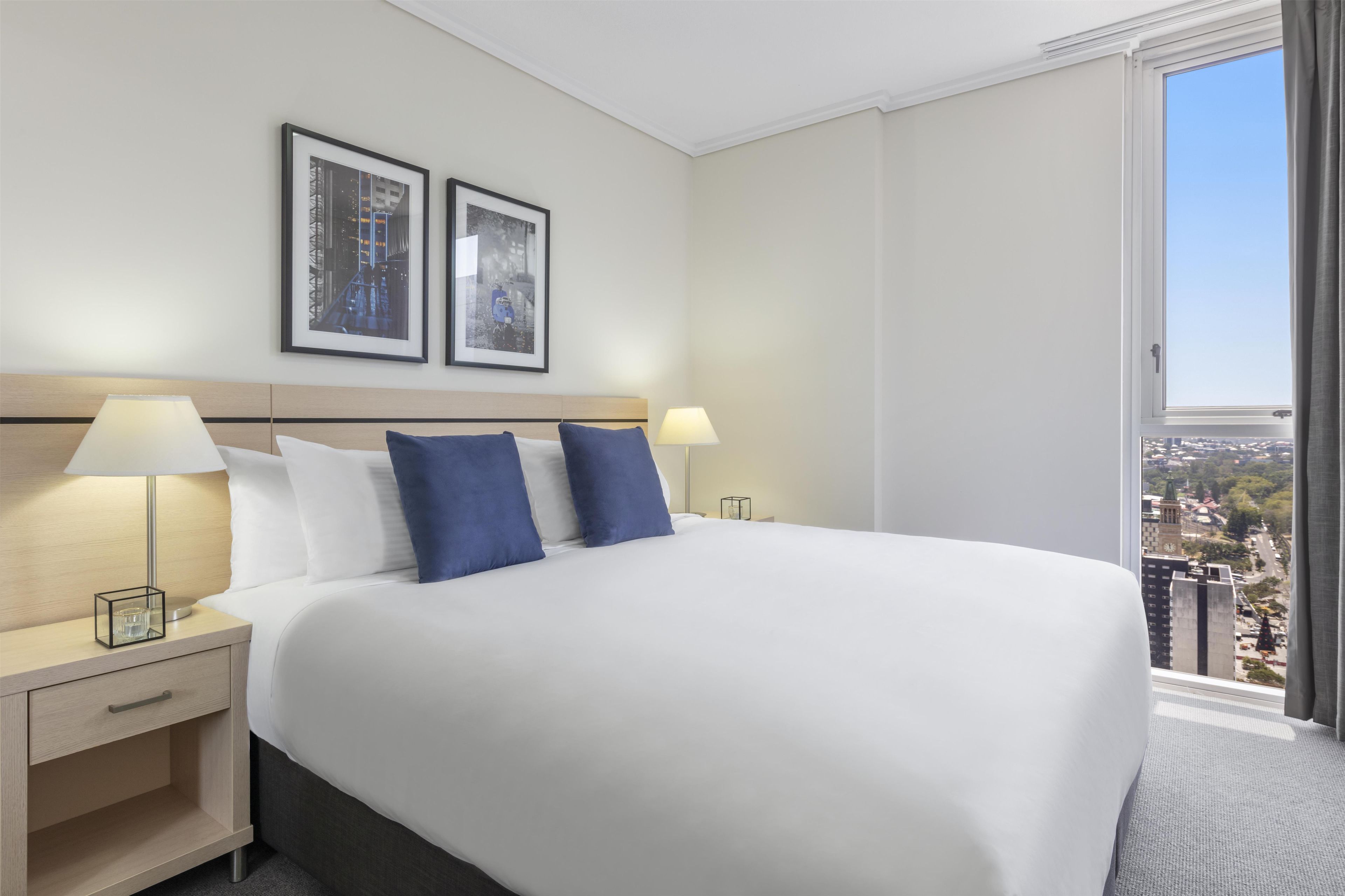 A hotel room with a large bed, two side tables with lamps, a nightstand, a drawer, and a view of the city through a large window.