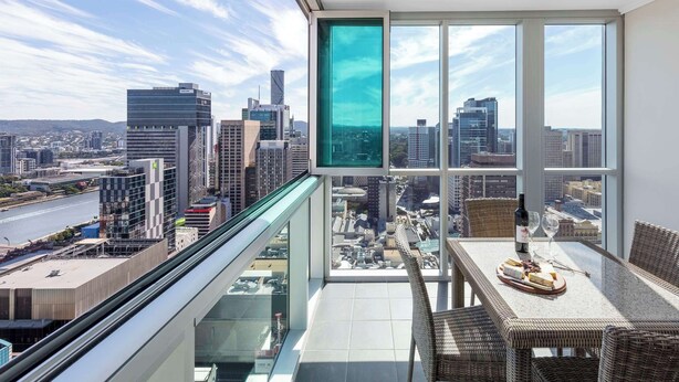 Un balcón con vistas a la ciudad, muebles de mimbre, una mesa con una botella y vasos, y un ventanal grande.