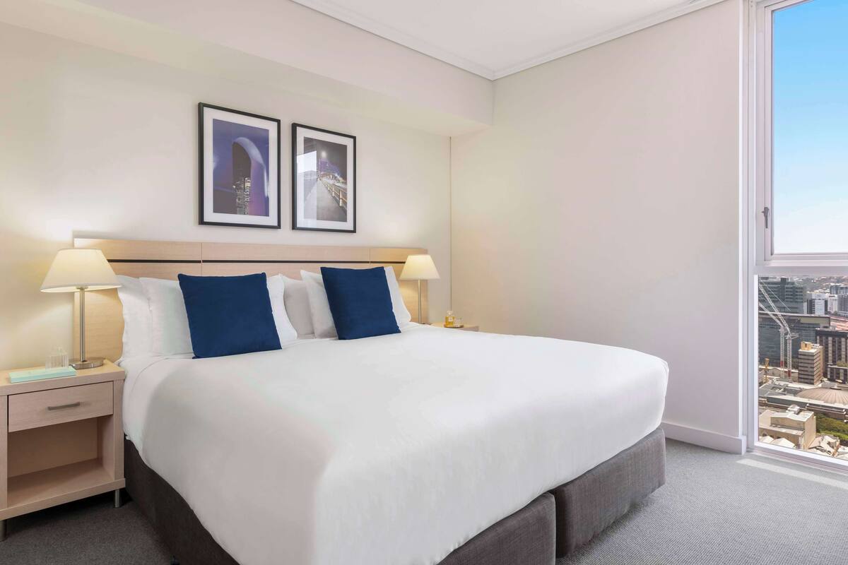 Ein Hotelzimmer mit einem großen Bett, zwei Nachttischen mit Lampen, einem kleinen Tischchen und Blick auf die Stadt durch ein großes Fenster.